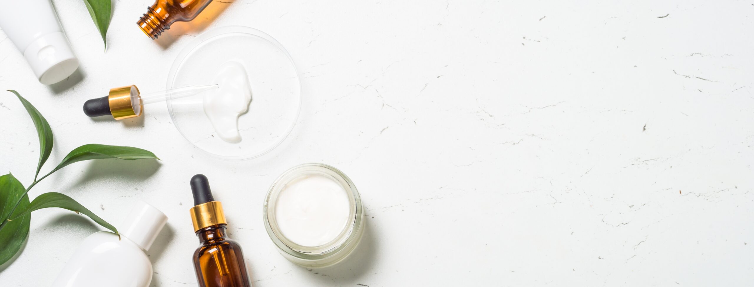 Serum bottles, a cream jar and green leaves on pale marble